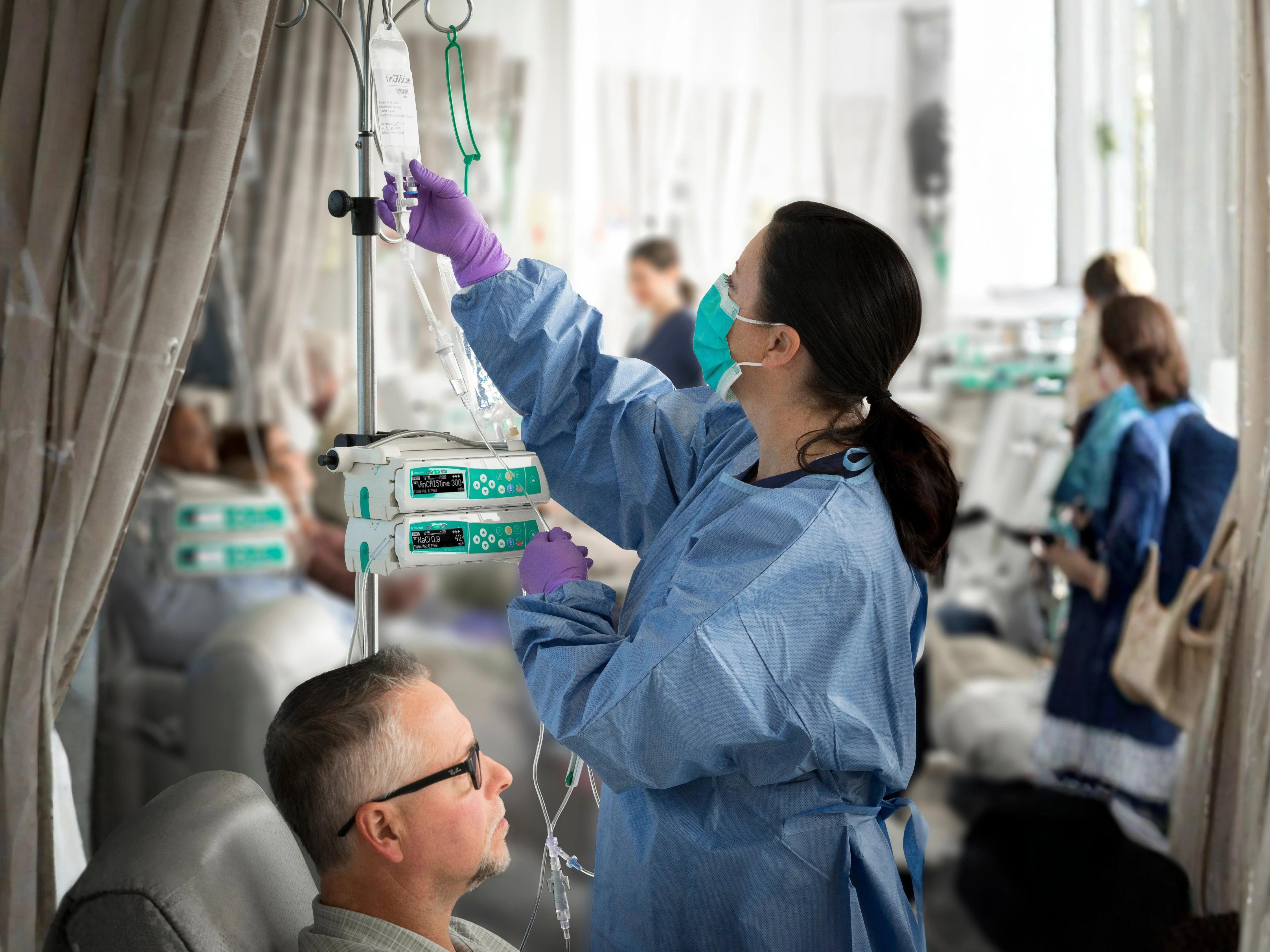 A clinician in protective clothing handling a suspended bag beside two stacked B. Braun Space pumps and a seated patient in a hospital setting.
