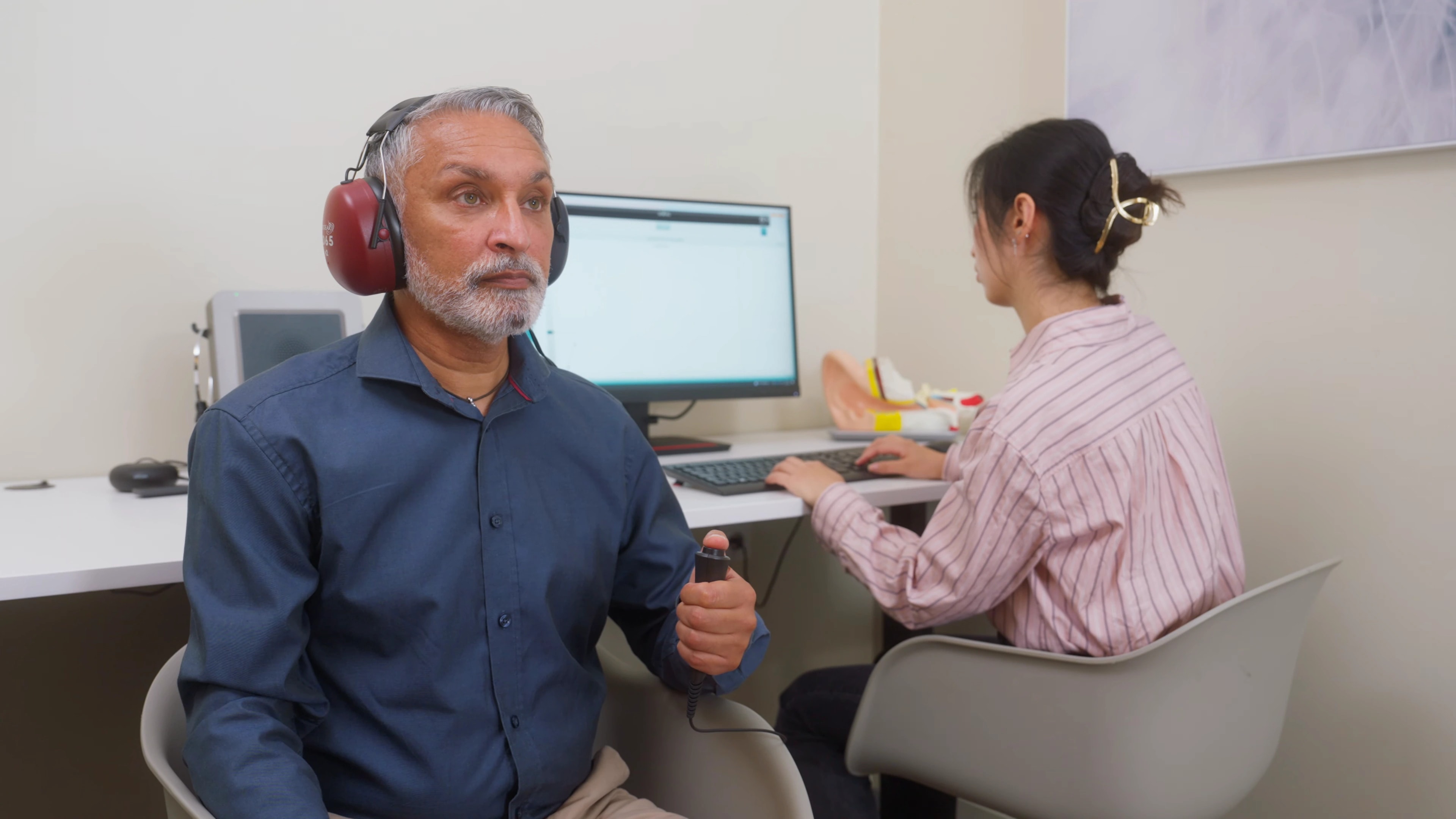 A person wearing audiometry headphones holds a response button while an operator runs an Interacoustics hearing assessment.